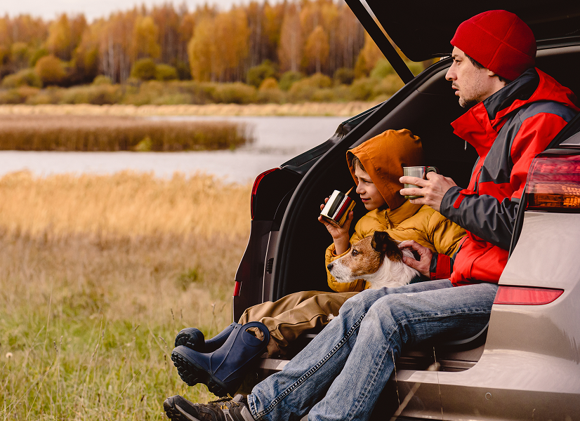 Familie mit Hund genießt Herbstblick Mann und Kind sitzen mit Hund im Kofferraum eines Autos, halten Tassen in der Hand und blicken auf den herbstlichen See und die Bäume.