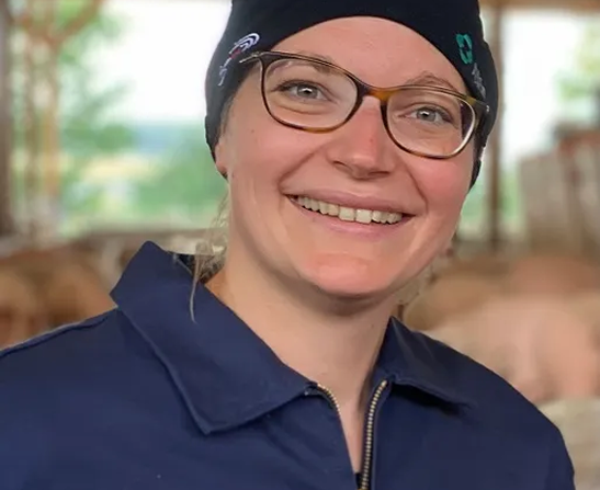 Porträt einer Mitarbeiterin in der Landwirtschaft Porträt einer lächelnden Frau mit Brille, dunkelblauer Arbeitskleidung und einer schwarzen Mütze vor einem Stallhintergrund.