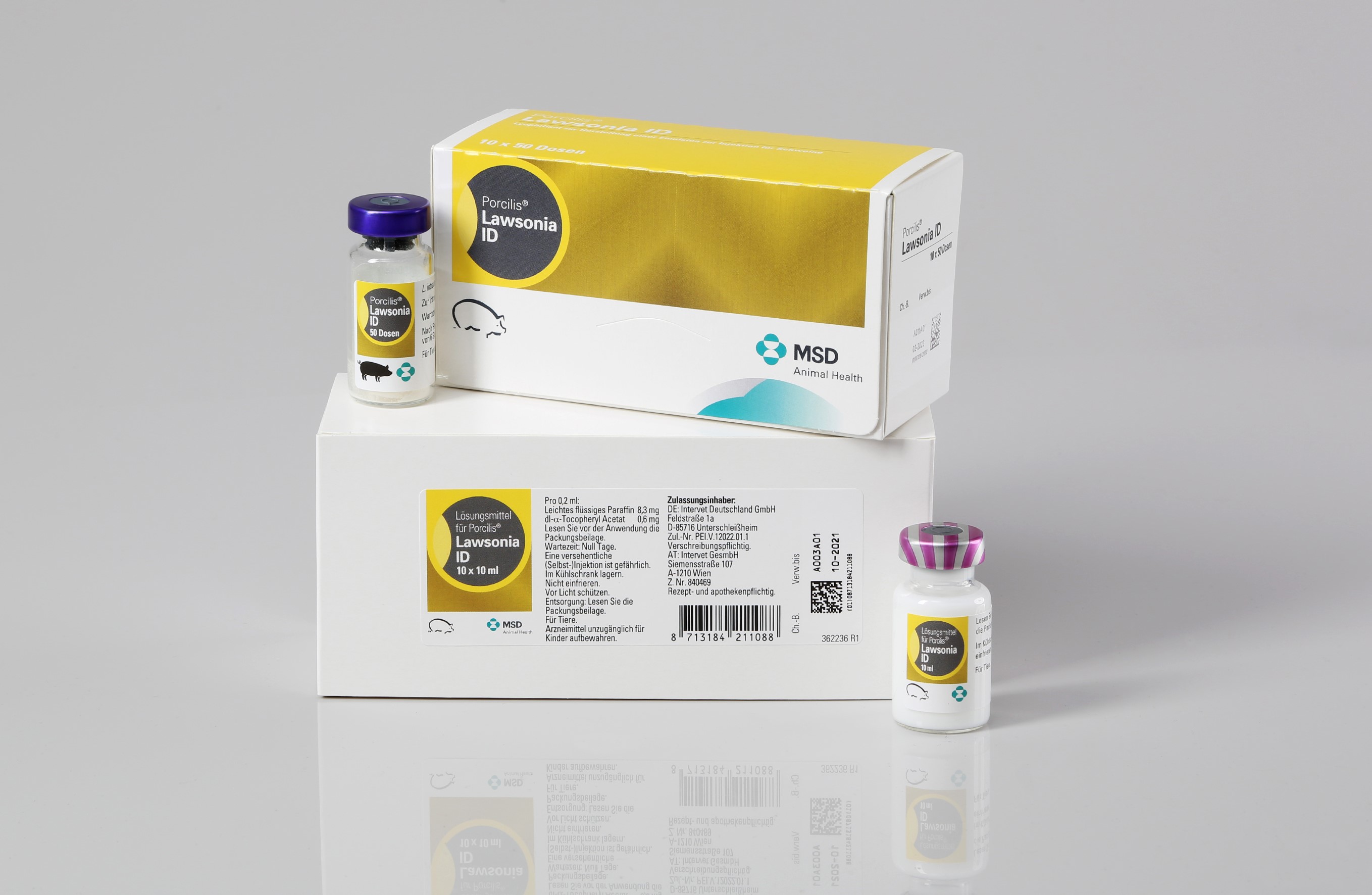 Packshot für einen Impfstoff. Eine Packung aus Karton steht auf einer weiteren Packung neben einem Glasfläschchen. Auf dem Boden steht ein weiteres Glasfläschchen mit einer weißen Flüssigkeit.